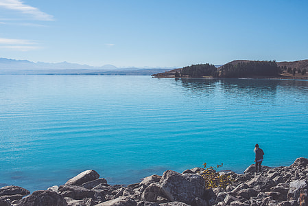 person standing on rocky ground near body of water under blue sky thumbnail