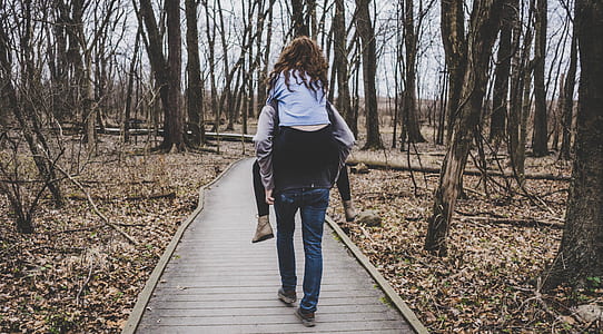 person piggy backing girl walking on wooden pathway between forest thumbnail