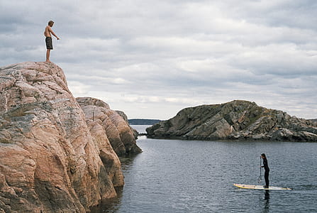 man standing on cliff near man riding surfboard thumbnail