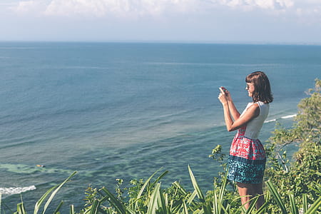 woman standing on ground filled with green grass holding smartphone taking photo of body of water thumbnail
