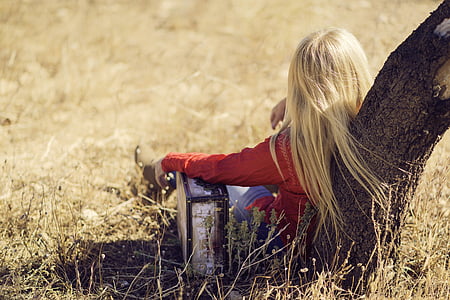 girl with blonde hair wearing red sweater and denim skinny pants leaning on brown tree on beige withered grass field during daytime thumbnail