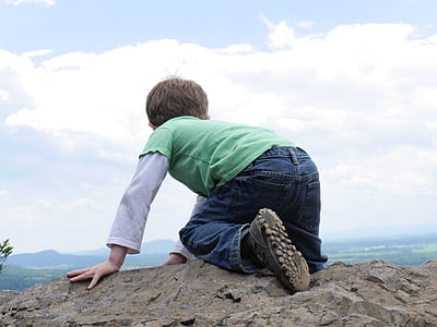 boy kneeling on cliff under white clouds during daytime thumbnail