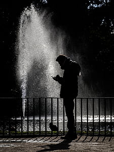 silhouette of man using phone near metal railing and fountain during daytime thumbnail