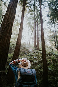person wearing blue long-sleeved top and brown hat standing beside tall trees during daytime thumbnail