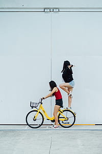 two women riding yellow step-through bicycle near white wall during daytime thumbnail