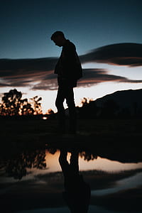 silhouette of man wearing jacket and pants walking beside body of water thumbnail
