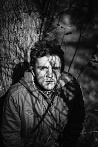 grayscale photo of man wearing zip-up jacket standing beside tree thumbnail