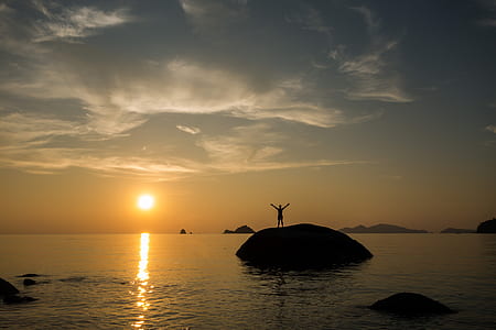 silhouette of man standing on stone near body of water during sunset thumbnail
