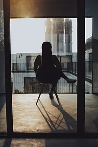 woman sitting on chair near window overlooking white building thumbnail