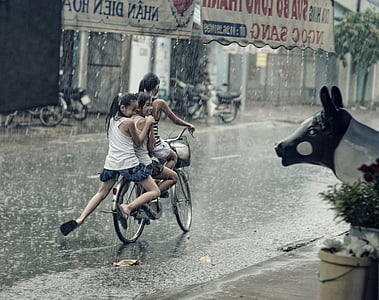 three girls riding bicycle traveling on road during rainy season thumbnail