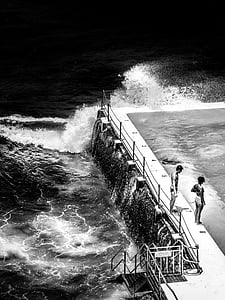 greyscale photo of two person standing on side of swimming pool near body of water thumbnail