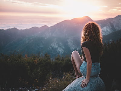 woman sitting on gray stone in front of white and black mountain at golden hour thumbnail