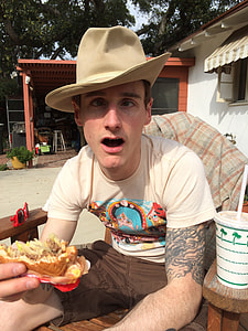 man in white shirt holding burger while sitting on brown armchair thumbnail