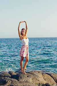 woman stands on rock formation raising her two hands beside body of water thumbnail