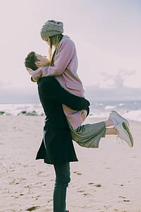 Man in Gray Coat Carrying Woman Wearing Pink Coat in Beach Near Shoreline and Body of Water thumbnail