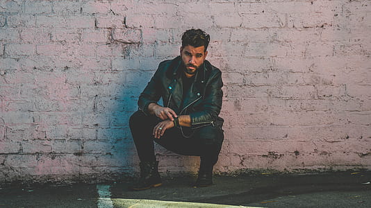 man wearing black jacket leaning on white wall thumbnail