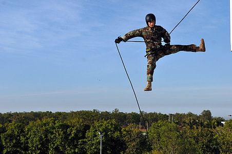soldier in mid-air wearing camouflage suit holding on rope below green leafed trees during daytime thumbnail