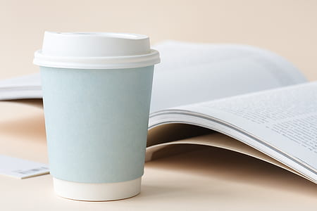 blue and white disposable coffee cup beside open book on table thumbnail