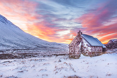 Brown House Surrounded by Snow Covered Field Near Snow Covered Mountain Under Yellow Blue and Orange Sunset thumbnail