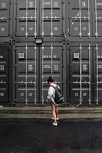 Photography of Woman in Gray Long Sleeve Shirt Carrying Black Backpack Standing in Front of Black Intermodal Container thumbnail