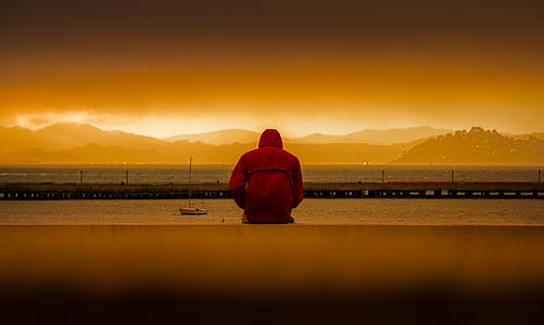Person Wearing Red Hoodie Sitting In Front of Body of Water thumbnail