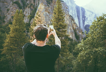 man wearing black t-shirt and holding phone while taking picture of mountain thumbnail