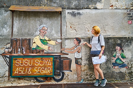 woman wearing gray standing behind gray wall with mural portraying man beside cart serving bowl to boy thumbnail
