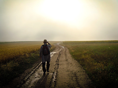 man in brown fedora hat and red backpack walking on path beside green field during daytime thumbnail