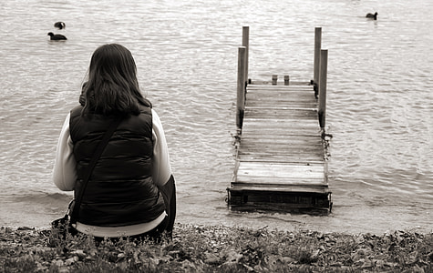 grayscale photo of woman in vest sitting on sand beside ocean thumbnail
