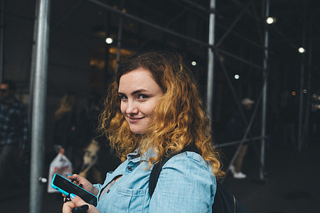 smiling woman wearing blue chambray button-up collared shirt holding blue smartphone near gray scaffolding tunnel thumbnail