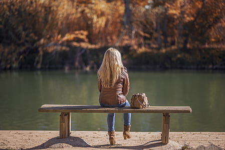 woman wearing brown leather jacket and blue jeans sitting on bench thumbnail