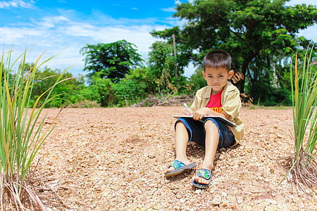 boy holding pen and book while sitting on ground in middle of trees thumbnail