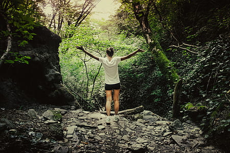 woman in white shirt and black shorts stands in the middle of forest thumbnail
