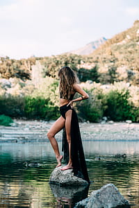 woman standing on rock surrounded by water near green leaf trees and mountain thumbnail