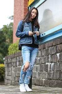 woman wearing denim jacket and jeans leaning on gray wall holding smartphone thumbnail