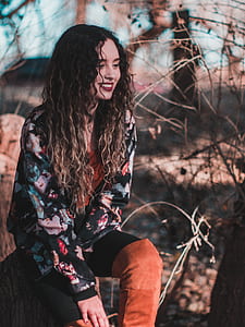 photo of smiling woman wearing black and multicolored cardigan sitting beside bare trees thumbnail