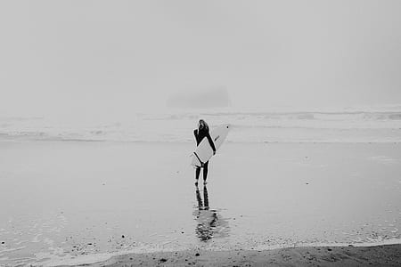 woman carrying surfboard while watching wave of beach rushing to the beach grayscale photography thumbnail