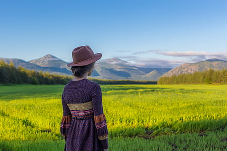 woman standing in front of green grass field and mountains under blue sky during daytime thumbnail