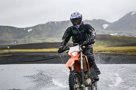 man riding orange dirt bike passing body of water during daytime thumbnail