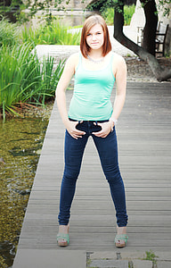 woman in teal tank top and blue jeans standing on brown wooden pathway near body of water thumbnail