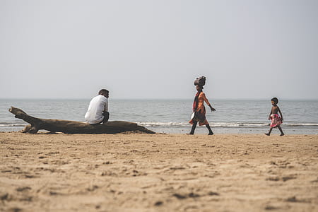man sitting on drifwood near woman and young girl walking towards him near ocean water during daytime thumbnail