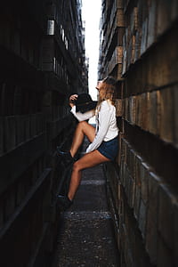 woman wearing white dress shirt and blue denim short shorts holding black cap stepping on brown wooden piled beams while leaning thumbnail