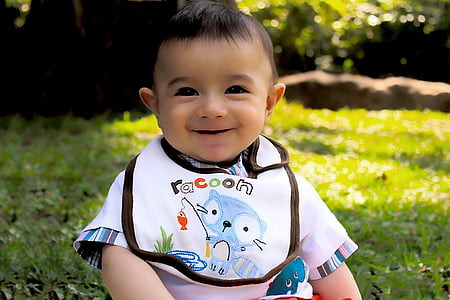 baby in white and multicolored shirt and Racoon bib sitting on green grass at daytime thumbnail