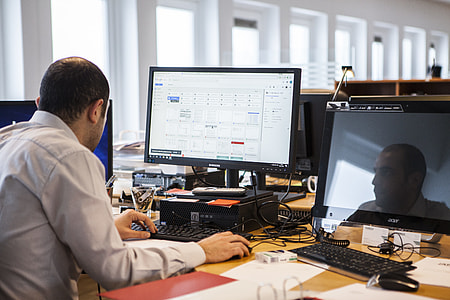 man in grey collared shirt sitting down facing flat screen computer monitor thumbnail