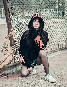 woman in black hoodie crouching near brown steel fence outdoor during daytime thumbnail