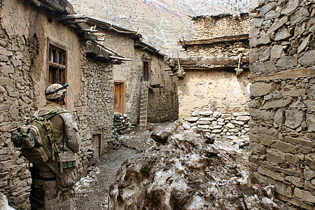 male soldier in gear set standing in front of rocky buildings thumbnail