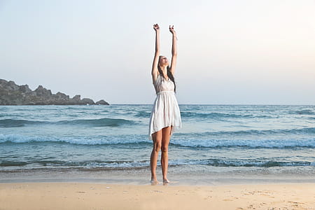 A young brunette woman wearing a white dress standing on the beach with her hands up thumbnail