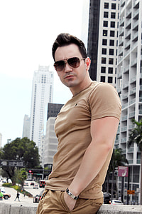 man wearing brown shirt and sunglasses standing on alley in front of building thumbnail