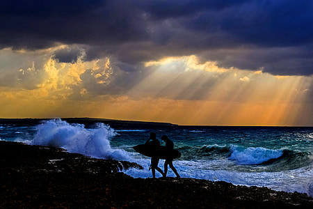 silhouette of two person on seashore holding surfboards near beach with waves under white clouds yellow skies sunset thumbnail