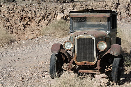 vintage brown and black car parked over dry ground with dried plants near rock formations thumbnail